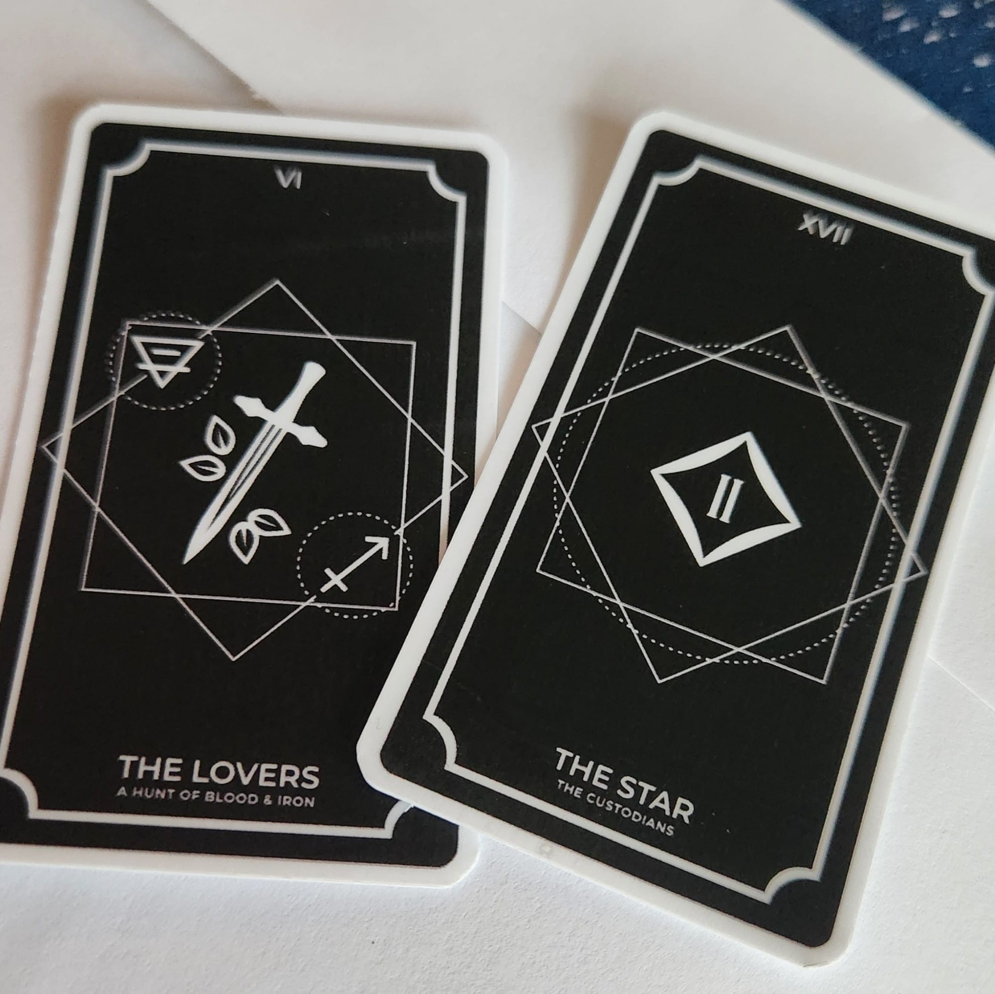 Tarot card stickers laying on top of an envelope. One is "The Lovers" with alchemic symbols around a sword in reference to A HUNT OF BLOOD & IRON. The other is "The Star" with a reference to the Spirits from THE CUSTODIANS series.